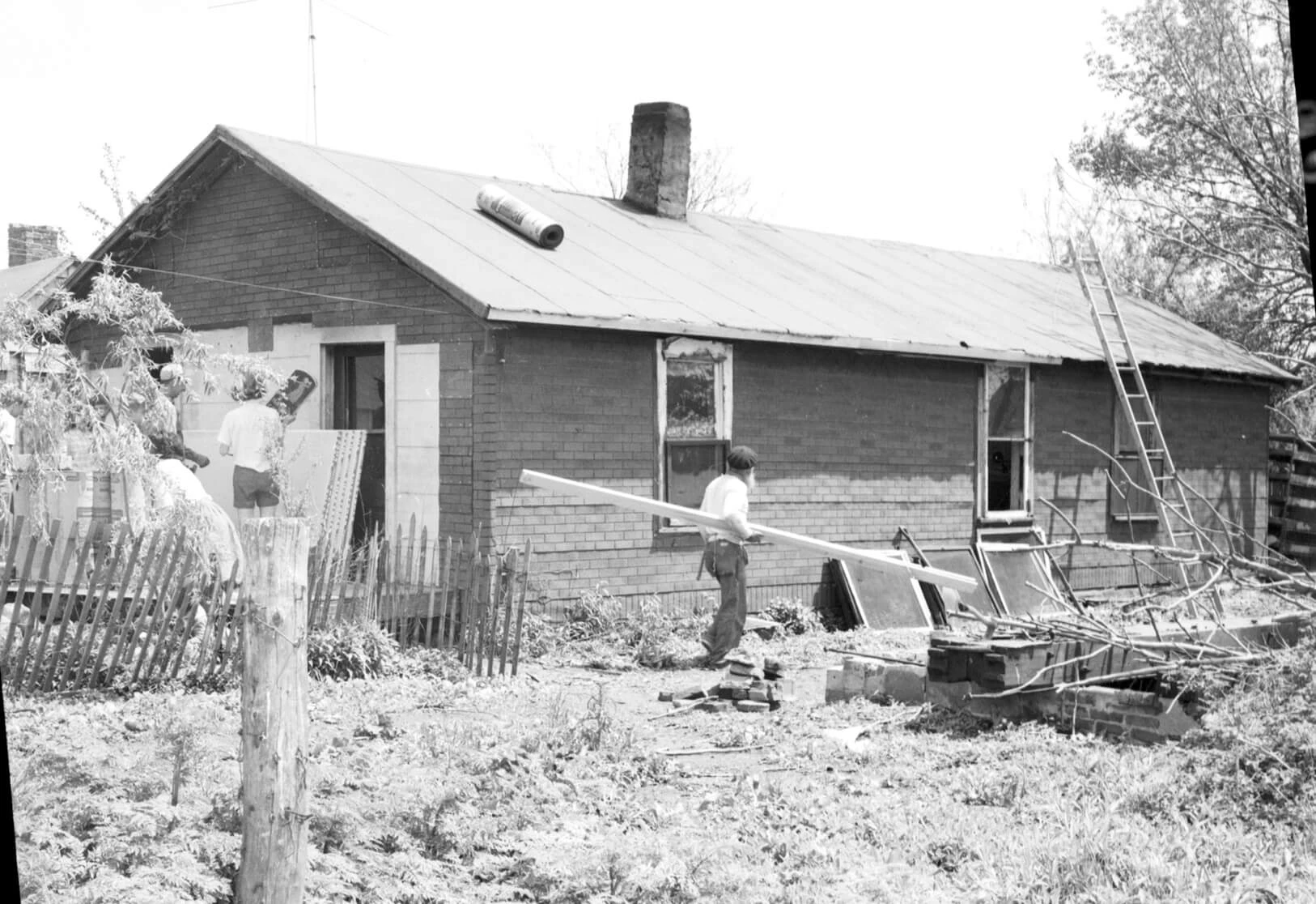 There is a small one-story home. A man is carrying a board toward a ladder which is leaning on the roof. A roll of roofing is on the shingle-less roof.