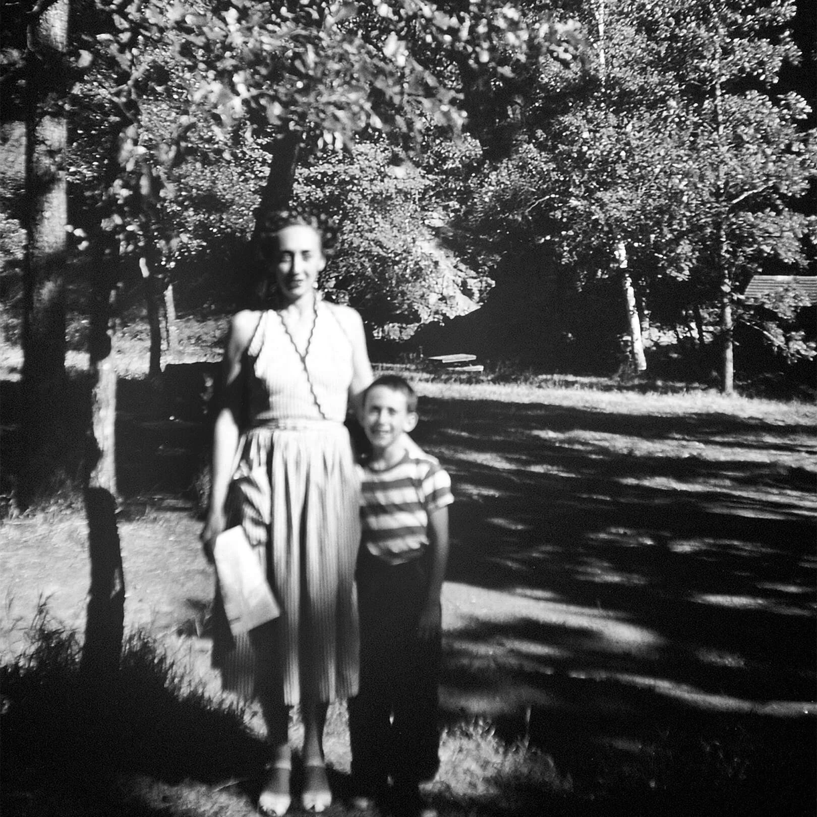 Black and white photo of a woman in a dress standing with a young boy. They are standing outside in a yard.