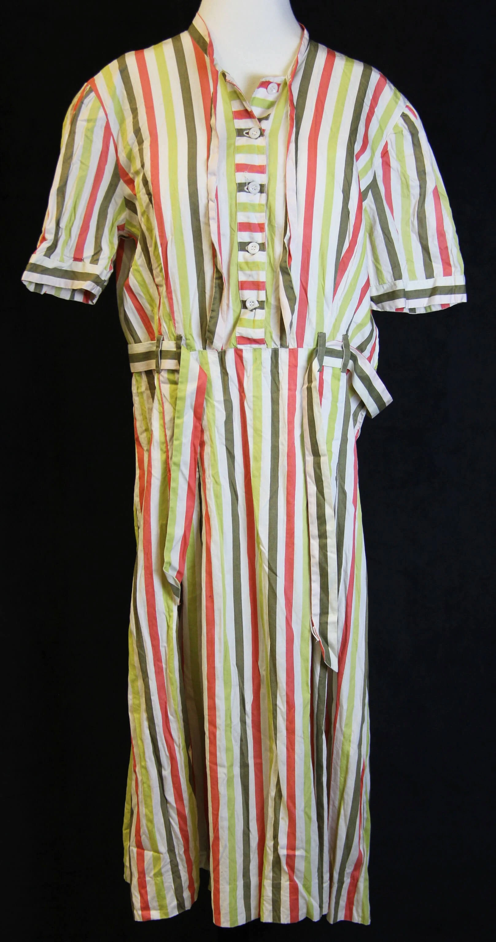 Color photo of a dress on a mannequin. The dress has white, light green, orange, and dark green vertical stripes of equal width. The dress has 5 buttons above the waist, a built in belt of the same material around the waist, and short sleeves.