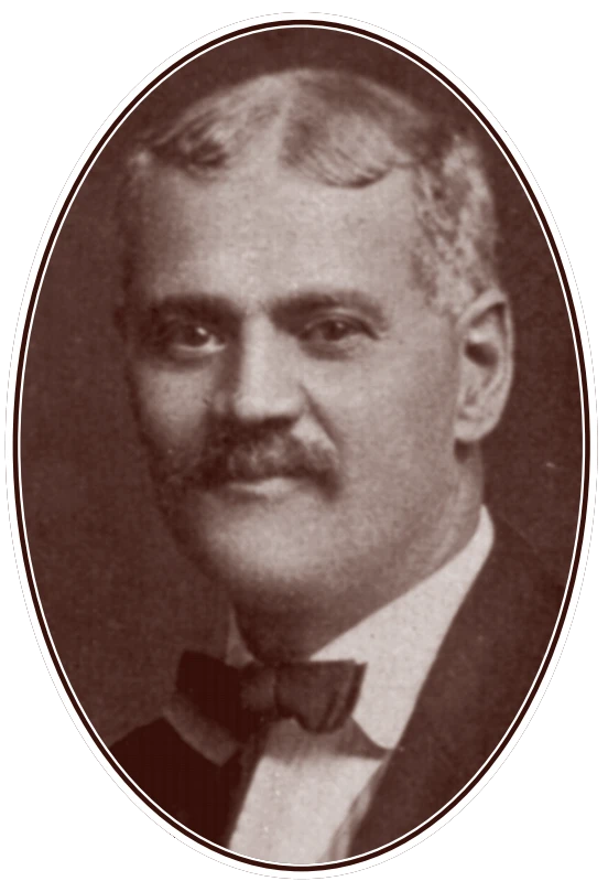 Picture of a man, shoulders up, wearing a suit and bow tie, with a mustache, looking directly into the camera.