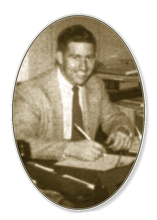 Grainy black and white photo of a light-skinned man in a light suit jacket and dark tie. He is sitting at a desk holding a pencil to paper. He is smiling and looking at the camera.
