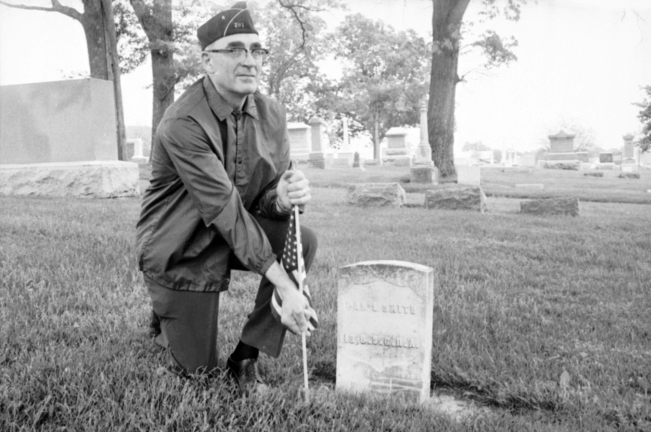 A man kneels, inserting an American flag next to a grave stone in a cemetery.